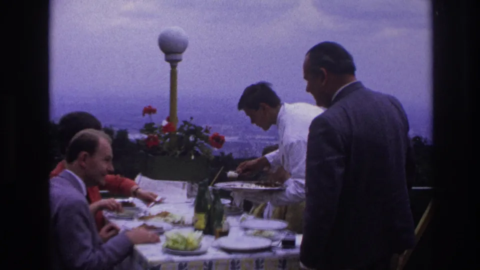 1967 home-movie frame of adults dining at a rooftop restaurant as a waiter in a white jacket serves food near flowers and a city view.