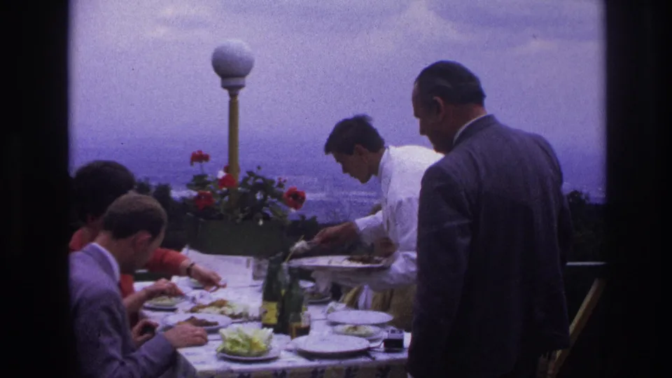 Home-movie view of a rooftop meal with adult diners, a waiter in white, patterned table linen, bottles, plates, flowers, and a city backdrop.