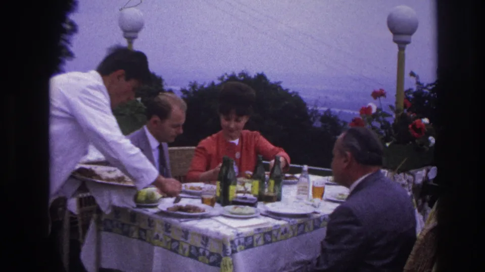 Adults gather around a rooftop dining table as a waiter leans in to serve, with bottled drinks, plates, red flowers, and a distant urban view.