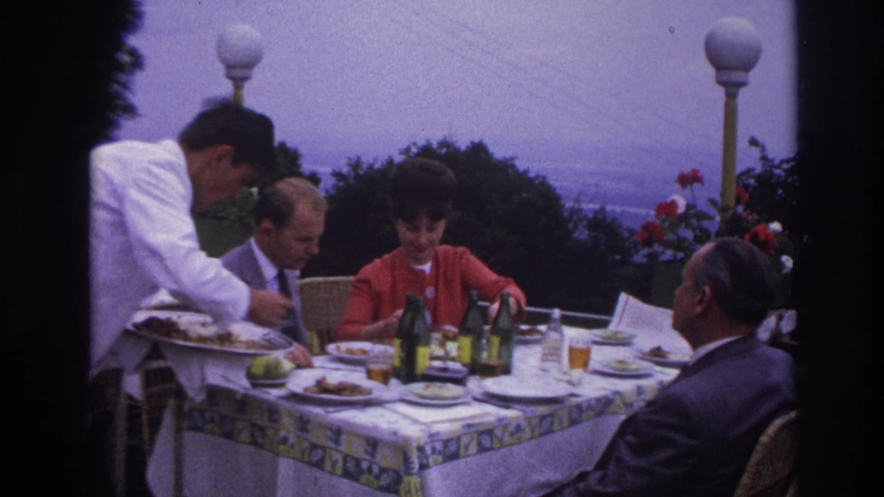 1967 France 8mm frame of adults at a terrace restaurant table while a waiter in a white jacket serves the meal.