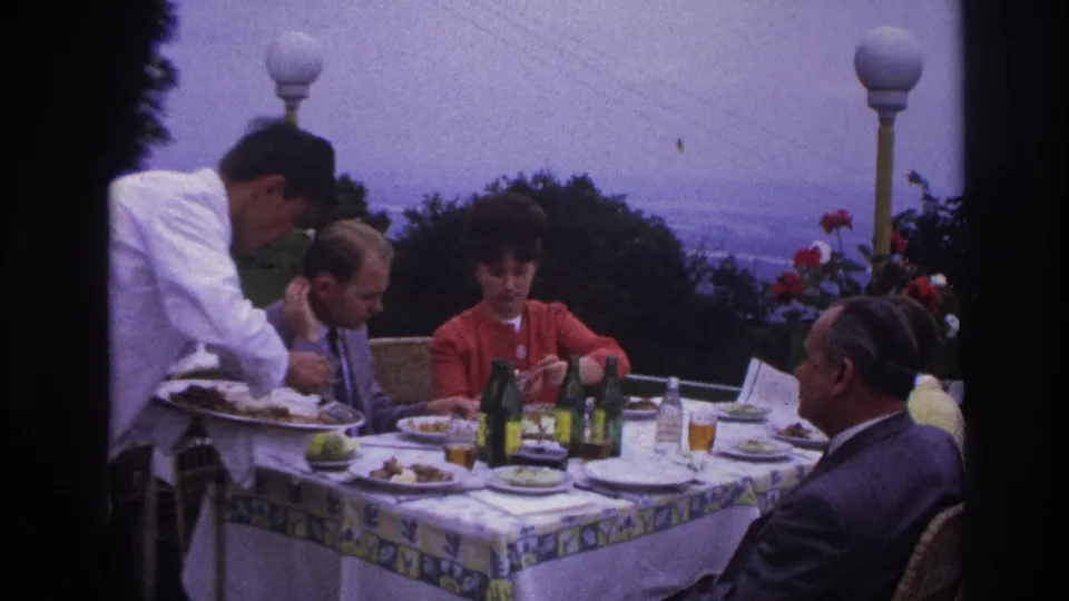 1967 France home-movie frame showing adults dining outdoors beside red flowers, with a hazy coastal landscape beyond the terrace.