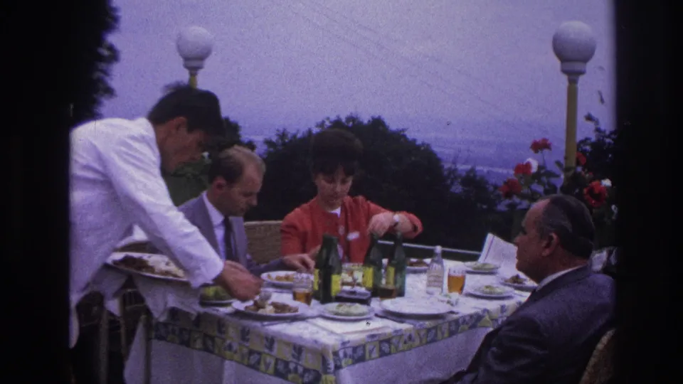 Vintage 8mm frame of an outdoor restaurant lunch in France, with four adults, a white-jacketed waiter, bottles, plates, and flowers.