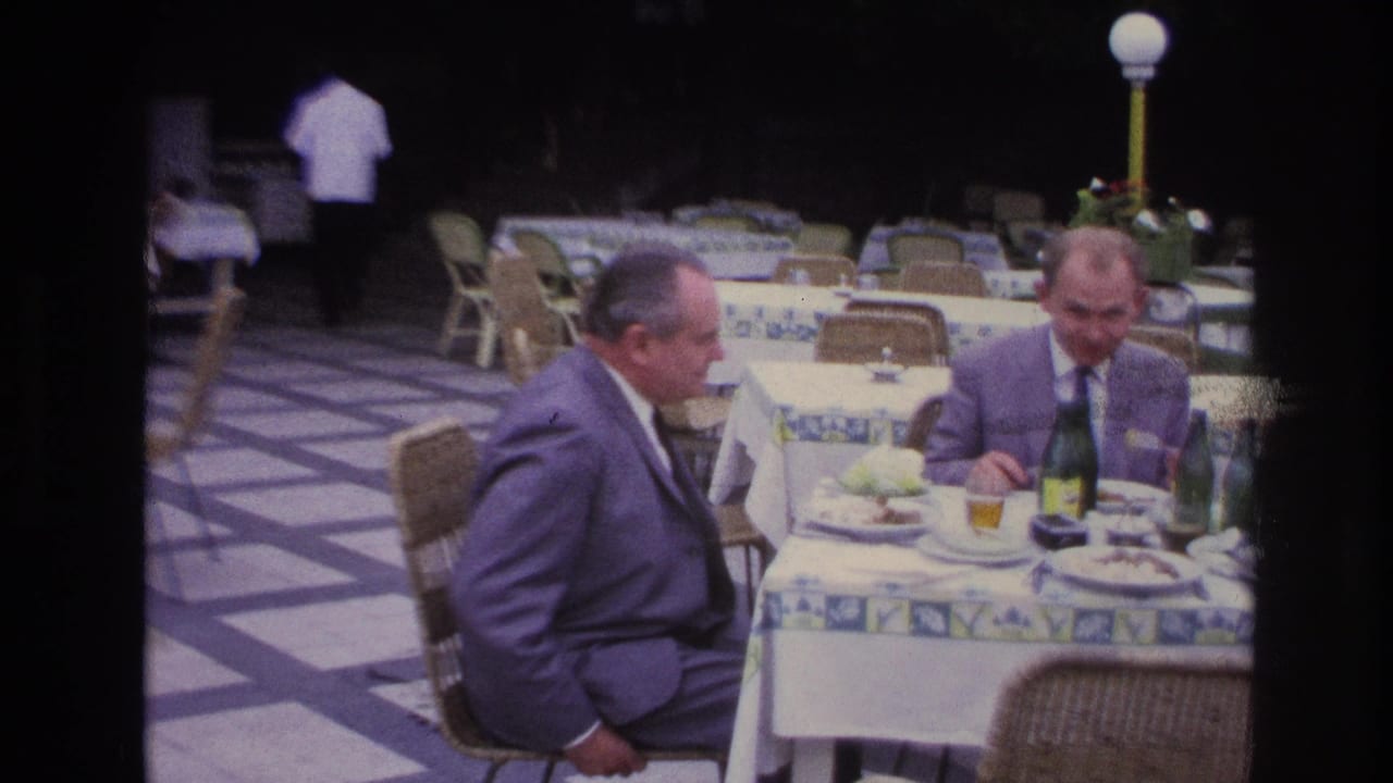 Two suited men dine at an outdoor restaurant table in France, with patterned tablecloths, wicker chairs, and drinks visible.