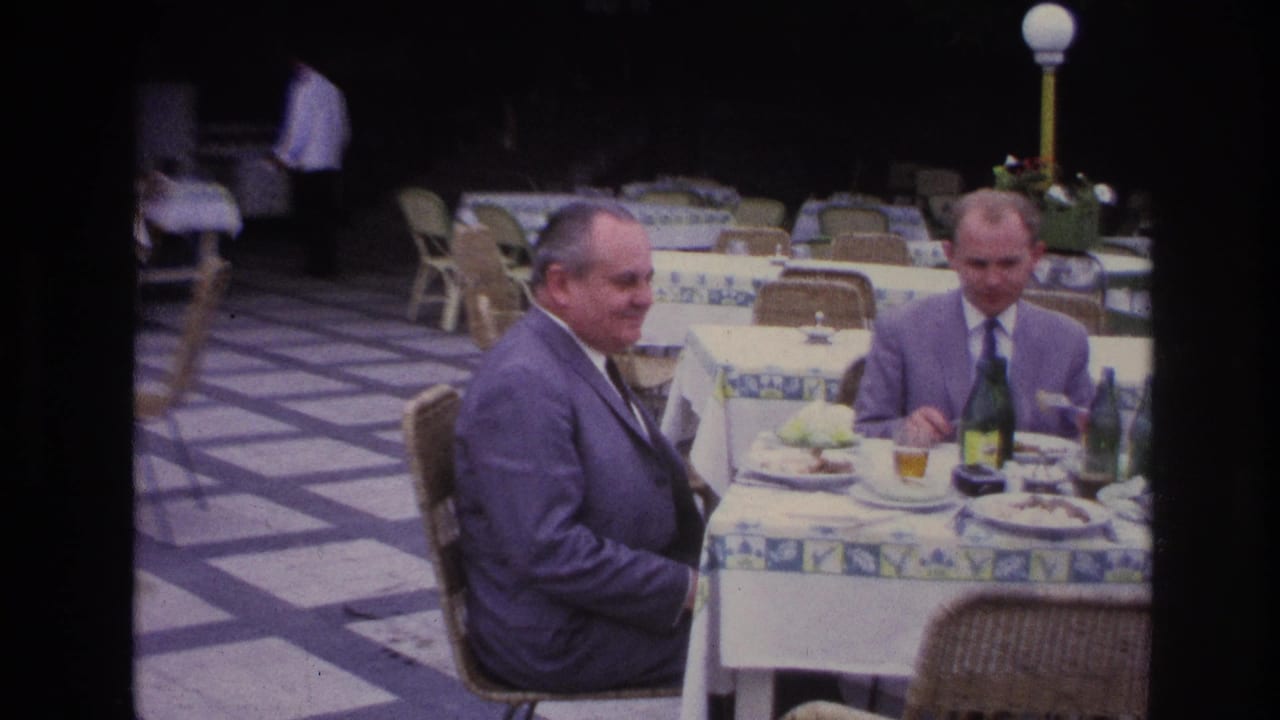 At an outdoor French restaurant, one suited man turns toward the camera while another remains seated by plates and bottles.