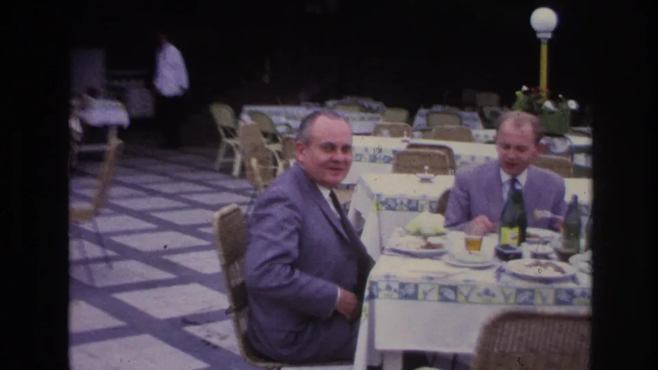 A suited man smiles toward the camera during an outdoor restaurant lunch in France, with another diner and table settings beside him.