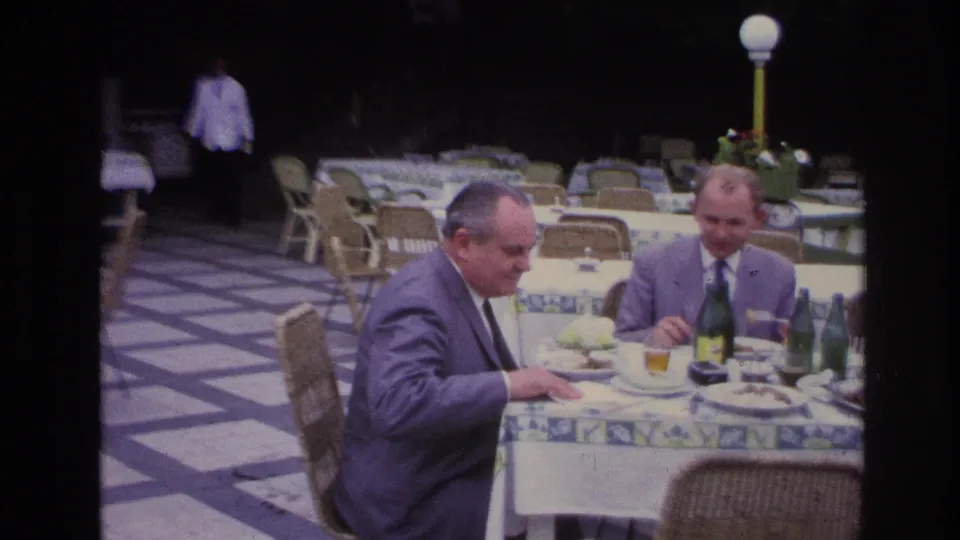 Two suited men continue eating at an outdoor restaurant table in France, with a waiter and rows of patio tables behind them.