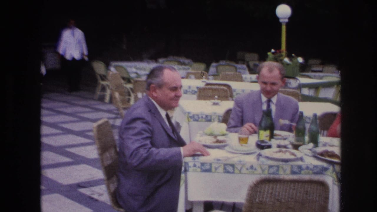 In 1967 France, two suited men dine at an outdoor restaurant table with patterned cloth, green bottles, plates, and patio seating.