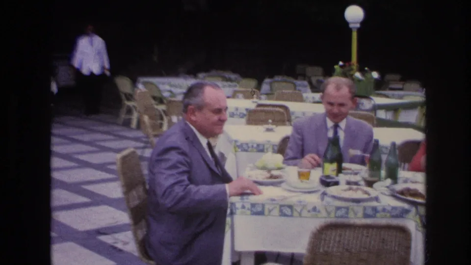 In 1967 France, two suited men dine at an outdoor restaurant table with patterned cloth, green bottles, plates, and patio seating.