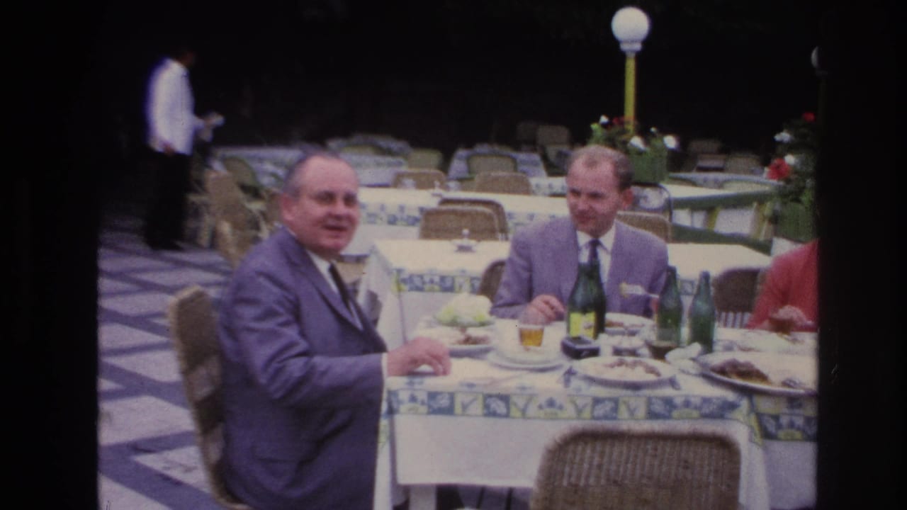 Two suited men face the camera while dining at an outdoor table with bottles and glasses.