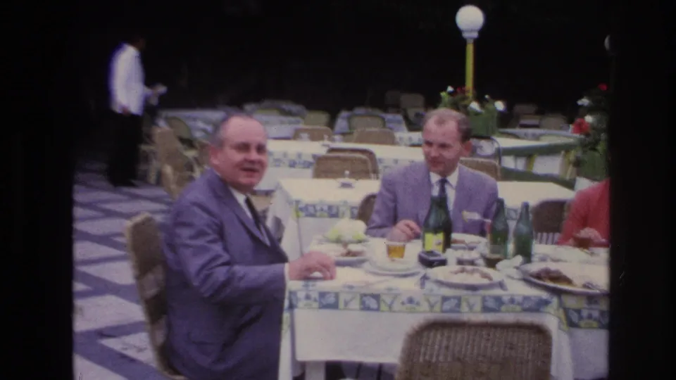 Two men in suits at a French outdoor restaurant look toward the camera while seated with plates, glasses, and green bottles on the table.
