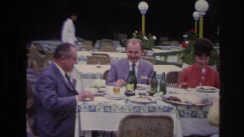 At a 1967 French outdoor restaurant, a woman in a red jacket joins two suited men at a table set with plates, bottles, and glasses.