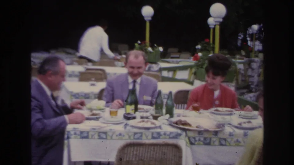 Three adults dine at a French patio restaurant, with a waiter in a white shirt moving behind the table and globe lamps visible nearby.