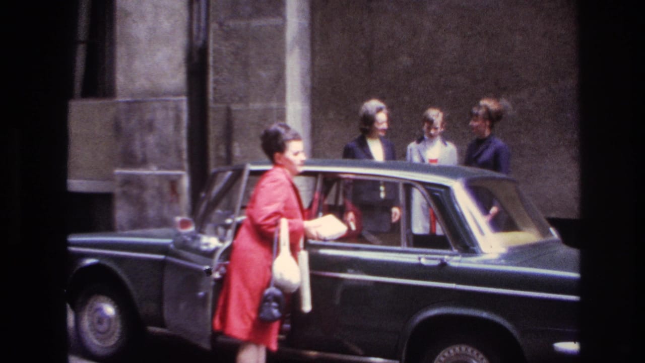 In 1967 France, a woman in a red coat approaches a dark sedan with a white handbag and booklet as three friends stand behind it.