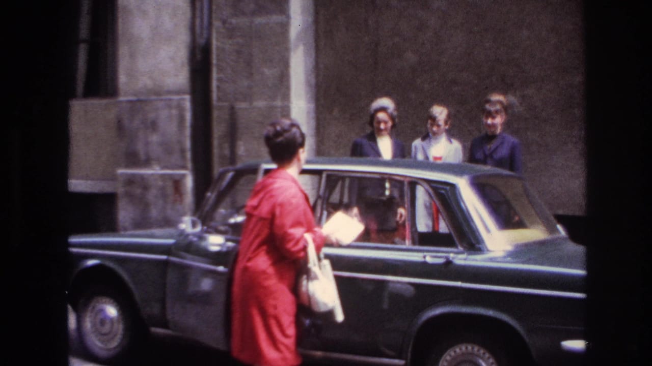 Woman in red coat reaches the rear door of a dark sedan with three people behind the car.