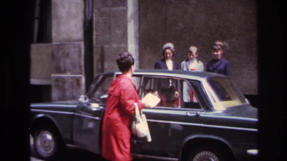 The woman in the red coat stands at the rear passenger door of the dark sedan, with three companions visible near the stone wall.