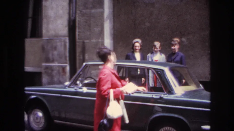 The woman turns back toward three friends while holding a booklet and standing beside the dark sedan on a French street.