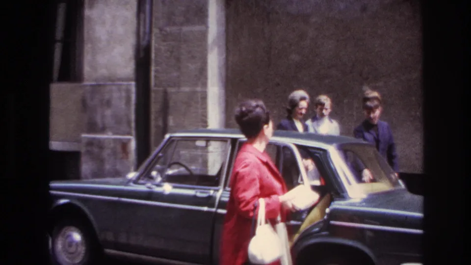 The rear passenger door is open on the dark sedan while the woman in a red coat faces three friends near the stone building.