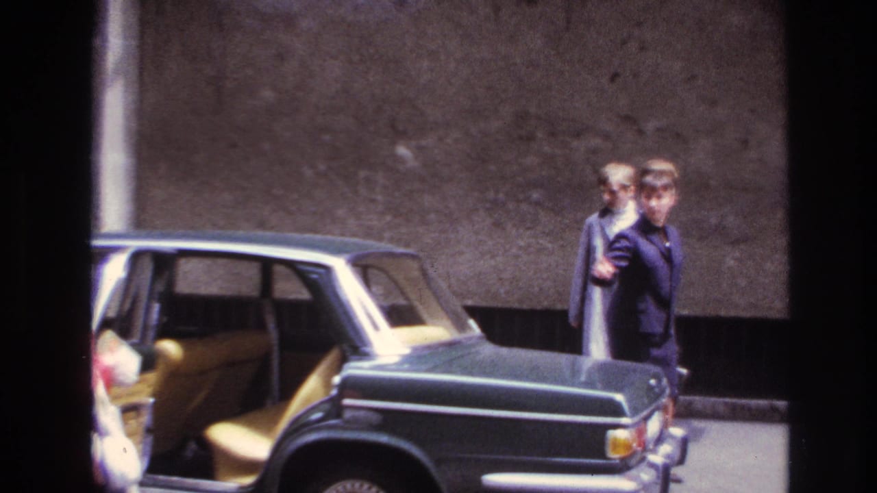 In 1967 France, two women in 1960s clothing stand by a dark green sedan with pale upholstery visible inside.