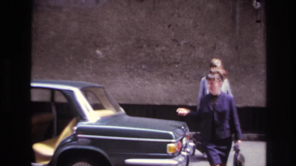 A woman in a dark suit walks near the open rear door of a classic sedan while another woman stands behind her.