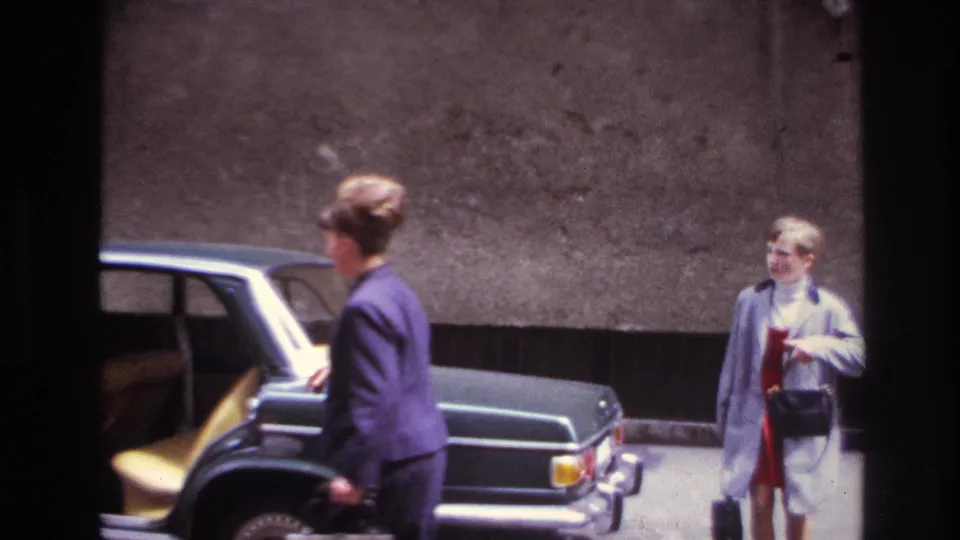 A woman in a dark suit walks past the sedan while a second woman in a light coat and red dress waits with a handbag.