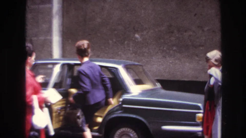 A woman in a dark suit boards the rear seat of a classic dark green sedan as others stand nearby in the street.