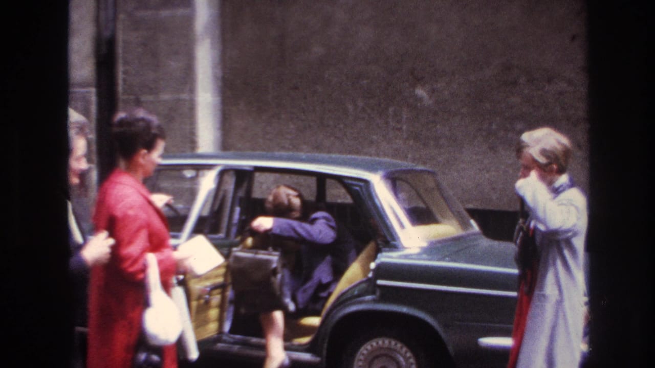 In France in 1967, adult women stand by a dark sedan while one person steps into the open rear door.