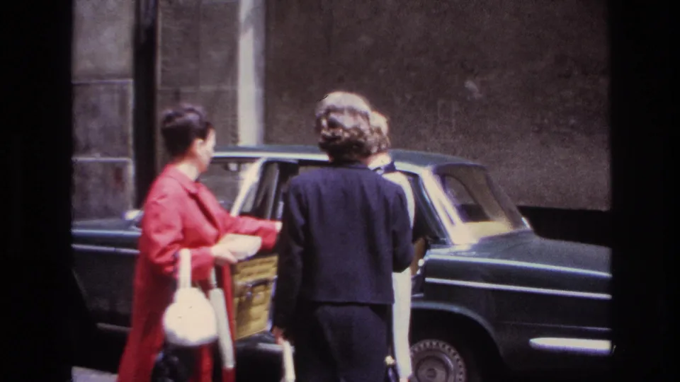 Adult women in formal coats stand beside the open rear door of a dark sedan on a French street in 1967.