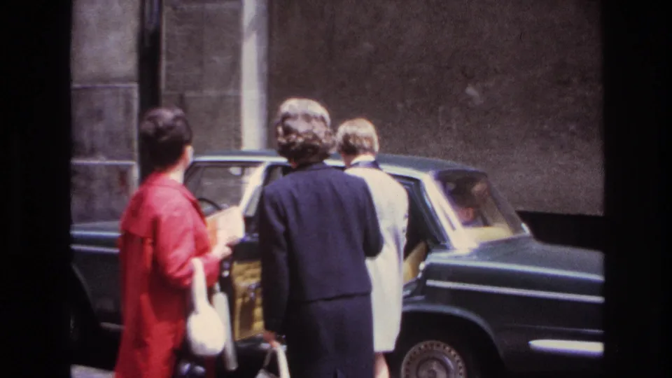 Women in dress coats face a dark sedan with yellow upholstery during a brief 1967 France home-movie street moment.