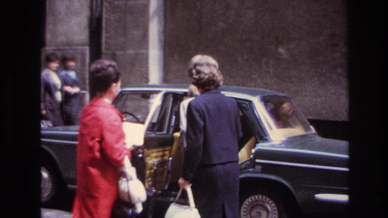 In 1967 France, two adult women stand by a dark sedan, with one in a red coat and another facing the open rear door.