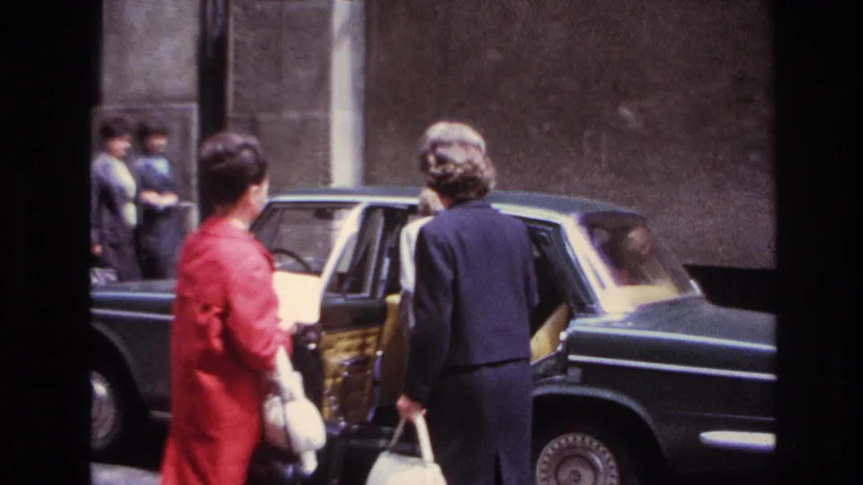 In 1967 France, two adult women stand by a dark sedan, with one in a red coat and another facing the open rear door.