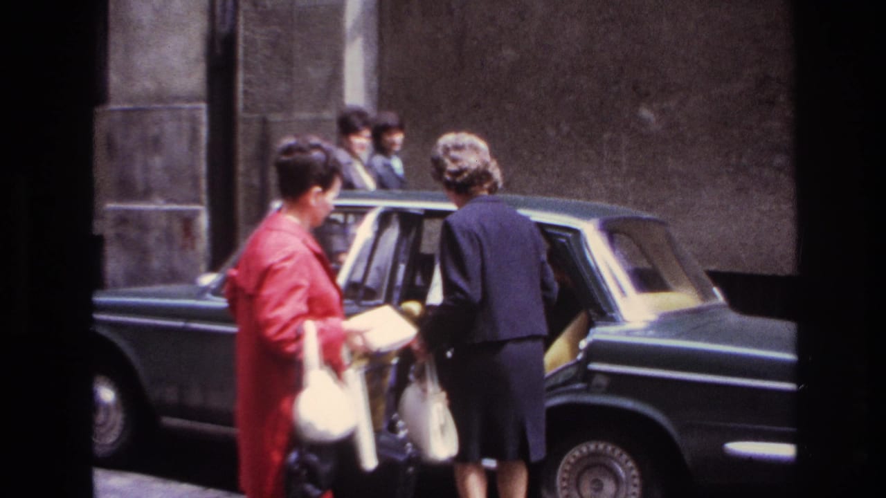 A woman leans into the rear seat while another woman waits beside the car.