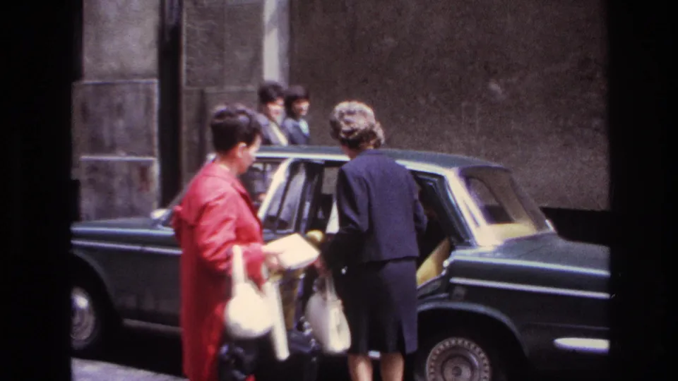 In 1967 France, a woman in a dark suit begins entering the rear seat of a dark sedan while a woman in a red coat stands nearby.