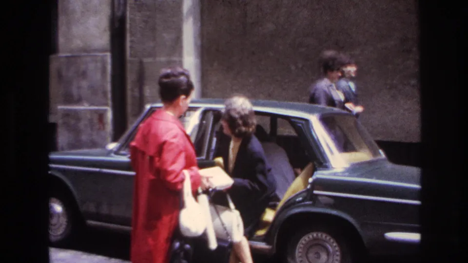 In 1967 France, one woman bends into the back seat of a dark sedan while the woman in the red coat observes from the curb.