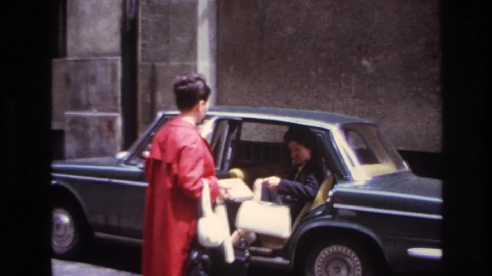 In 1967 France, the rear passenger is seated inside the dark sedan while the woman in the red coat stands beside the open door.