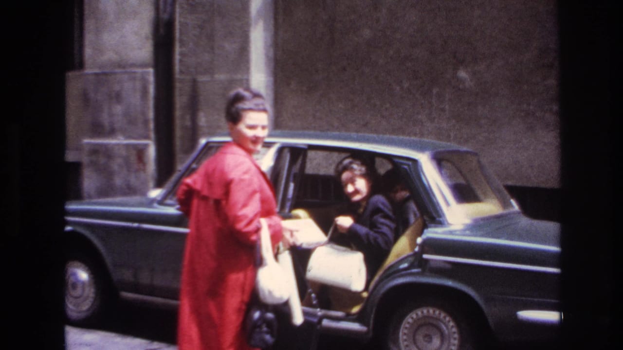 In 1967 France, a woman in a red coat and white handbag helps a woman in dark clothing settle into a dark sedan.