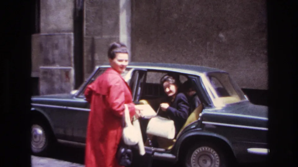 In 1967 France, a woman in a red coat and white handbag helps a woman in dark clothing settle into a dark sedan.