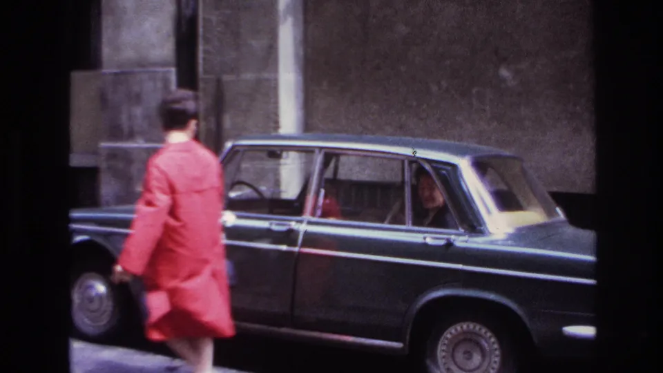 The woman in the red coat walks away along the street while a passenger remains visible through the sedan window.