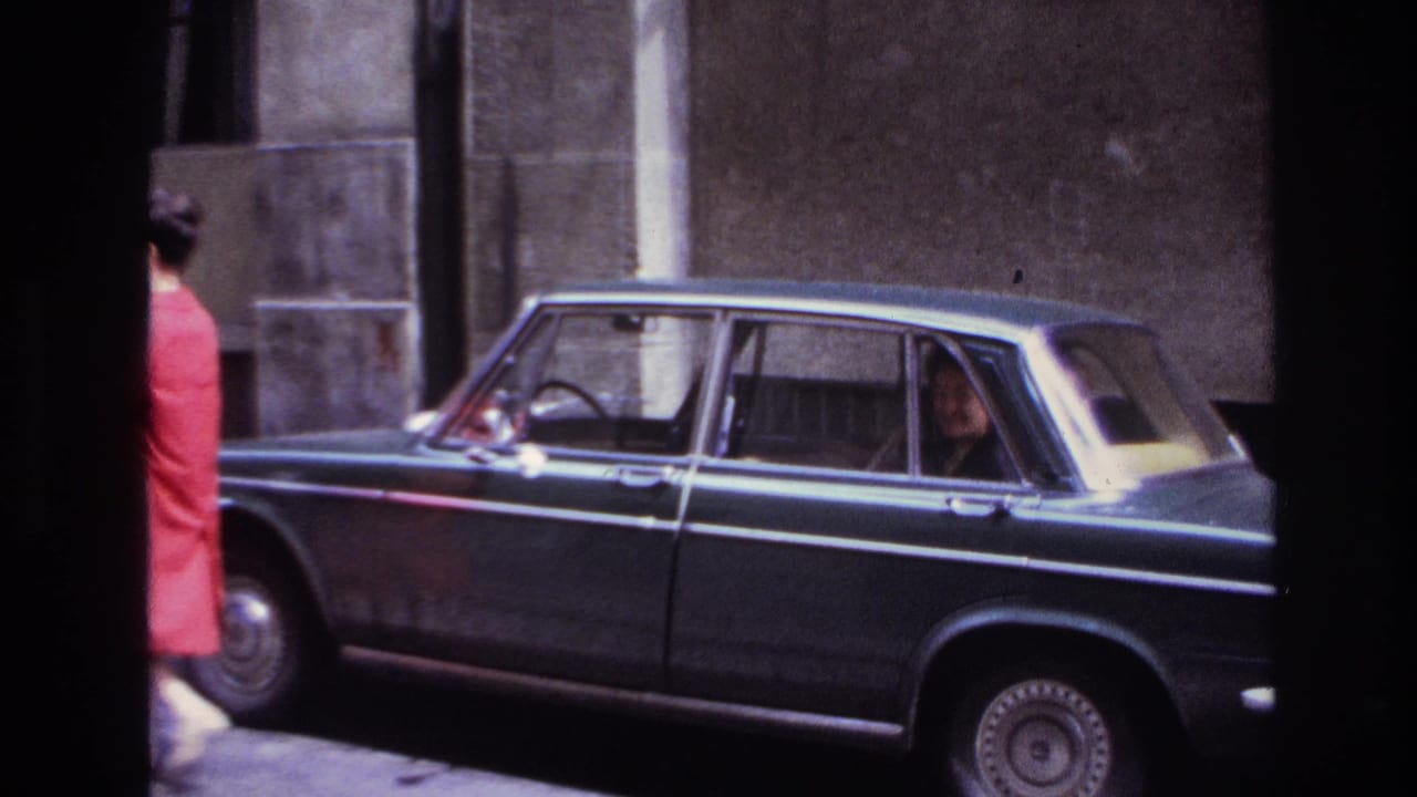 1967 France home-movie frame showing a woman in red near a dark four-door sedan with chrome trim and seated occupants.