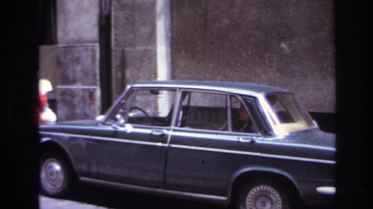 Dark sedan parked by a stone wall with occupants visible through the windows.