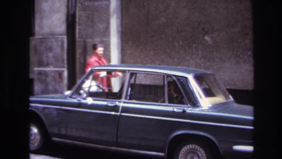 1967 France frame with the woman in red partly behind the sedan, with stone building details and car occupants visible.