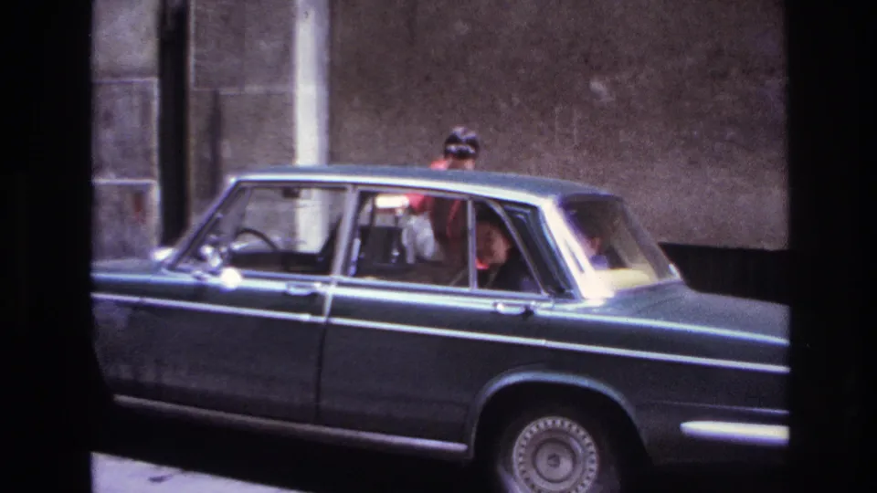 1967 France home-movie frame showing a woman at the side of a dark sedan while passengers sit inside.