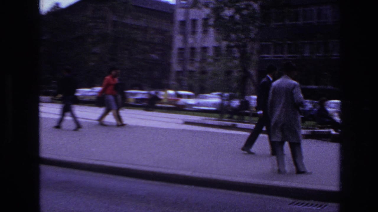 1967 France street scene with pedestrians crossing near parked cars and multi-story buildings, seen through dark film-frame edges.