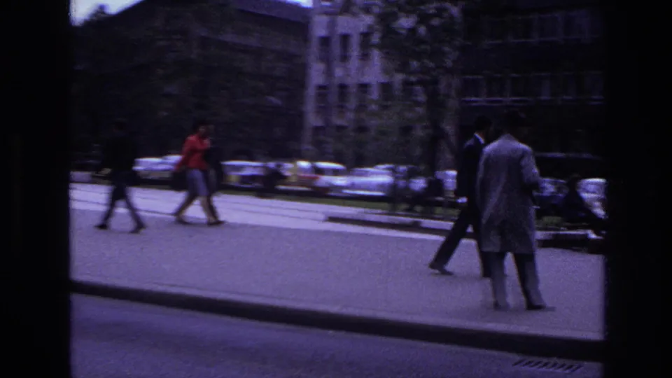 1967 France street scene with pedestrians crossing near parked cars and multi-story buildings, seen through dark film-frame edges.