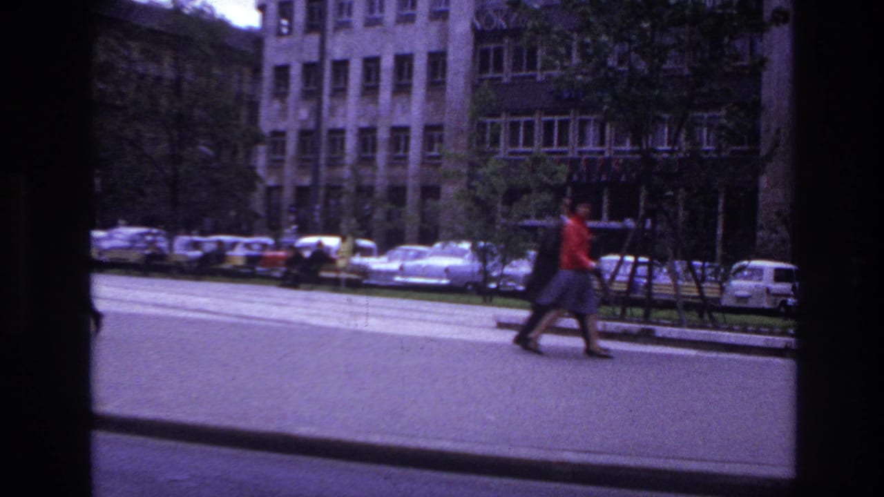 1967 France boulevard view with a pedestrian in red passing parked cars, trees, and tall urban buildings.