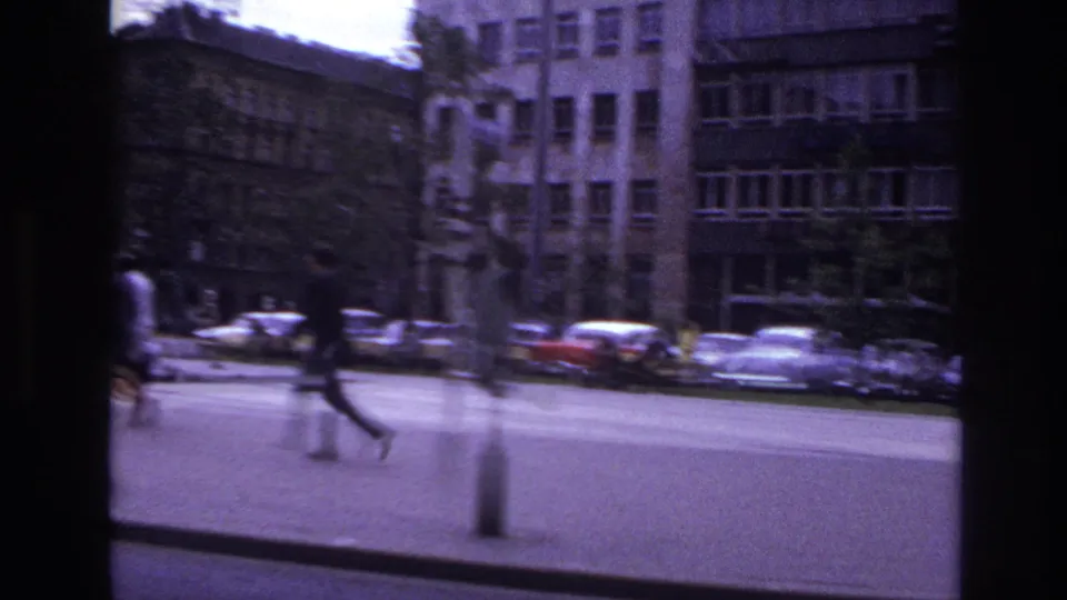 1967 France street footage showing a pedestrian with a light bag moving across a broad sidewalk near parked cars and urban facades.