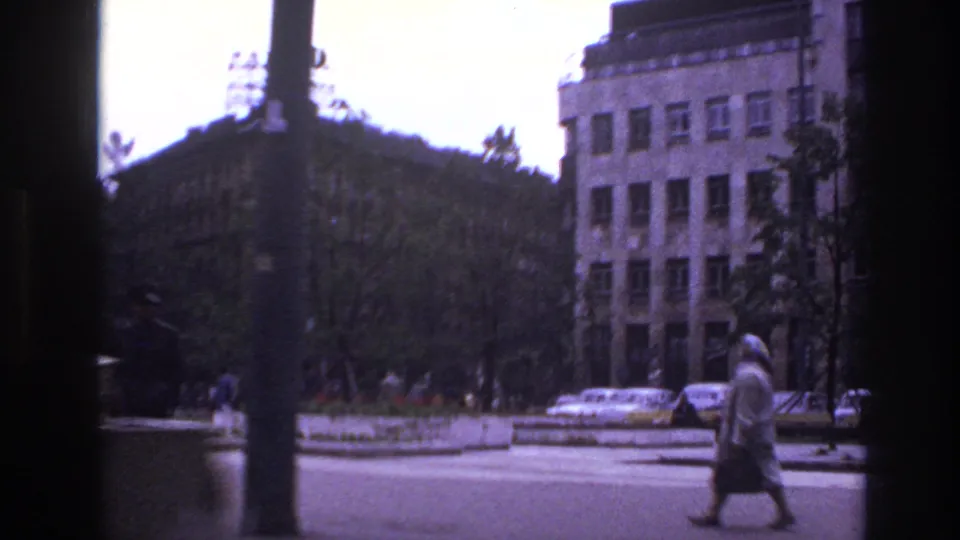 1967 France urban sidewalk view with a woman walking past parked cars, trees, and tall buildings in soft 8mm color.