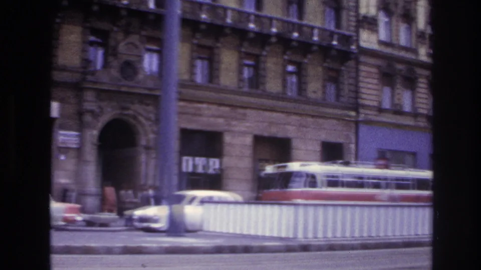 1967 France street scene with a tram, cars, pedestrians, large arched doorway, shop signage, and pale construction-style railing.