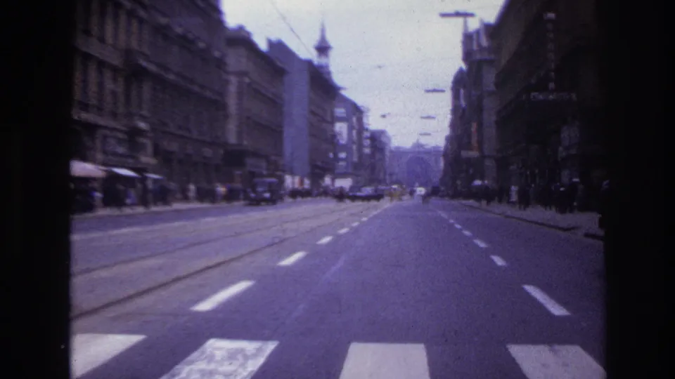 Moving-vehicle view along a wide French city street in 1967, showing tram rails, crosswalk stripes, shopfronts, older vehicles, and dense facades.
