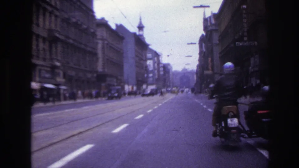 A motorbike rider appears at the curb of a 1967 France city street, with tram tracks, cars, shopfronts, and tall facades extending into the distance.