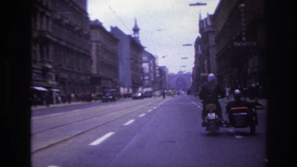 1967 France street view from a moving vehicle, with tram tracks, pedestrians, older buildings, cars, and a motorcycle with sidecar in front.